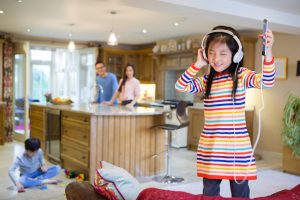 Little girl listening to music in the family kitchen. Her parents are looking and smiling at her, while her brother plays with his toys on the floor.